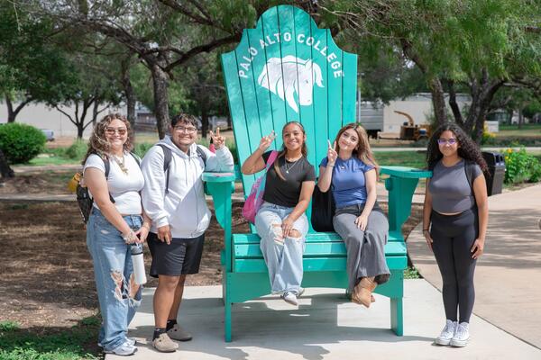 Three students stand around a comically large turquoise beach chair, while two students sit on the chair.