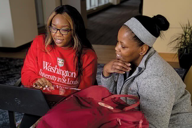 Two women wearing Washington University merchandise work on a laptop. 