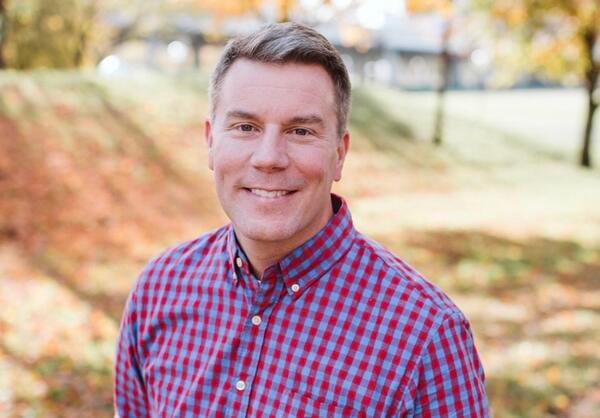Matt Bergman smiles for a headshot outside in a red plaid shirt