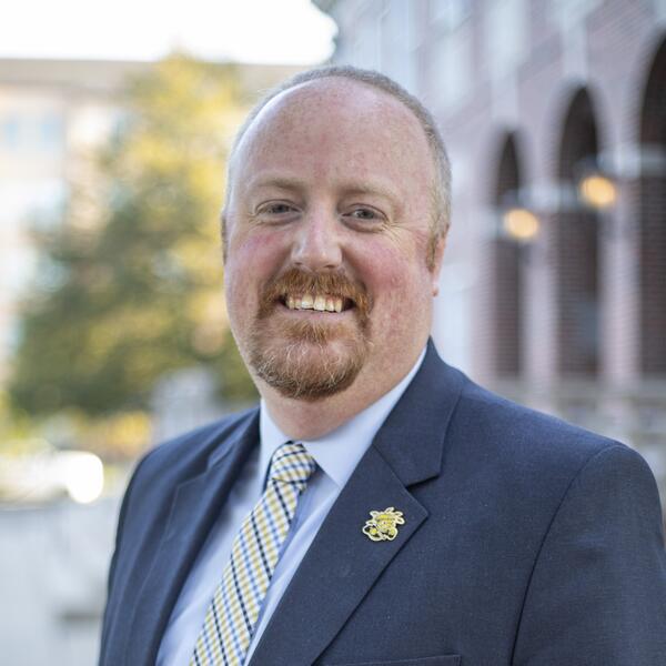 Brett Bruner smiles for a headshot outside. He’s wearing a navy suitcoat, a lighter blue shirt and a patterned yellow tie.