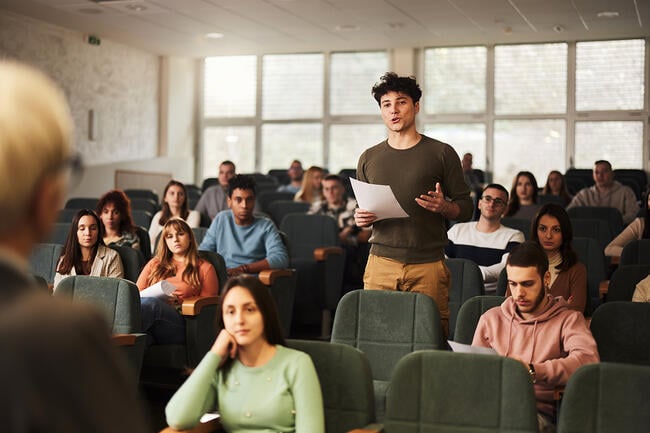 Smiling college student standing in a class during his oral exam in an amphitheater.