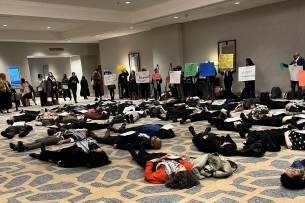 A photograph of a "die-in" protest at the Modern Language Association annual convention. Dozens of people are lying on the floor in a hotel, and people are standing around them with signs.