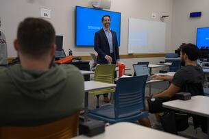 An instructor stands in front of a classroom, two students are visible in the foreground, facing the instructor