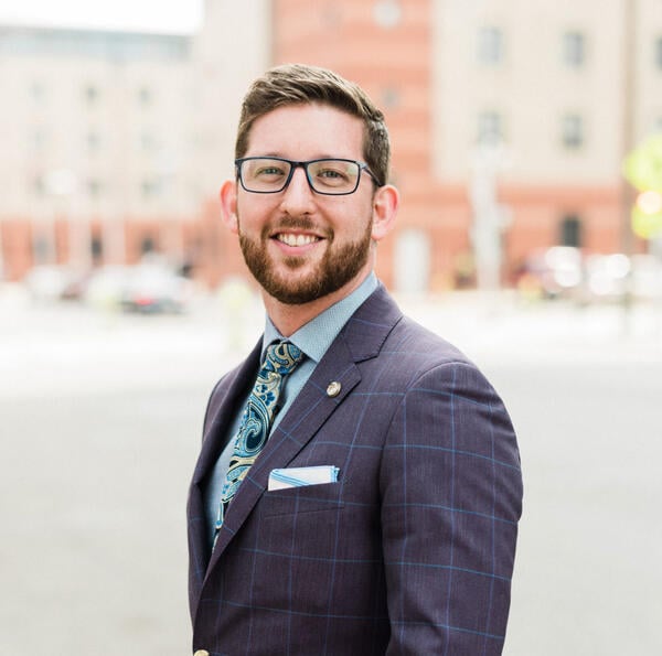 Eddy Conroy smiles for a headshot in an urban setting wearing a gray suite coat, blue shirt and patterned blue tie. He has short brown hair, black eyeglasses and a neat beard. 