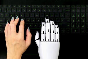 Human hand and a robot hand both hover over a computer keyboard. 