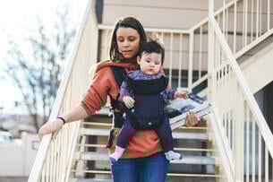 Millennial mother university student walking down housing stairs holding books with infant in a front carrier.