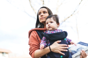 A woman is carrying a baby on her chest in a fabric carrier and holds a textbook in her left hand