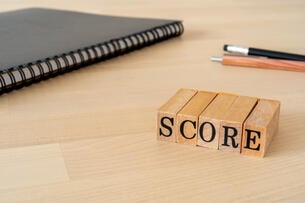 The word "Score" is spelled out in wooden stamps, on a table next to a black spiral notebook and two pens.
