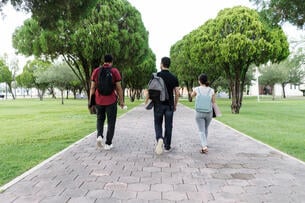 A photo of three Latinx college students walking away from a camera down a campus walkway, each wearing backpacks.