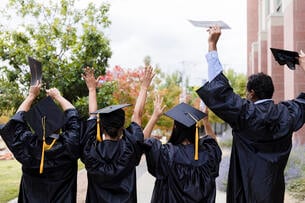A rear view of four unrecognizable graduates cheering after their graduation. The hold up their hands, their caps and their diplomas
