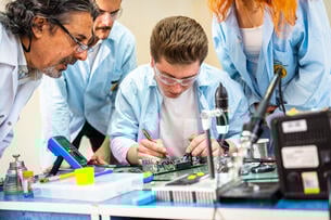An instructor and two students lean over a student in protective goggles working.
