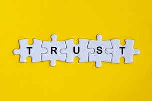Five jigsaw puzzle pieces spelling the word "TRUST" are linked together against a yellow background.