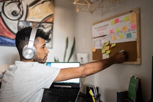 A young man wearing headphones adjusts his corkboard to update his planner