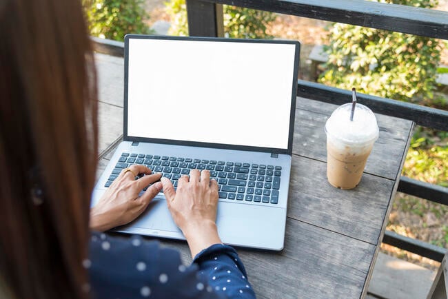 A close-up photo of a woman's hands typing on a laptop. She works outdoors -- plants can be seen in the background -- and a coffee drink is on the table next to the laptop.