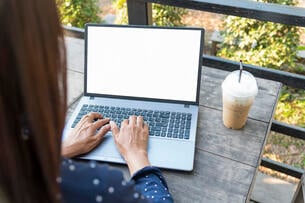 A close-up photo of a woman's hands typing on a laptop. She works outdoors -- plants can be seen in the background -- and a coffee drink is on the table next to the laptop.