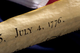 A stock photo of an aged copy of the Declaration of Independence with the words "July 4, 1776" visible, against an apparent American flag background.