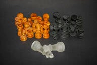 Groupings of brown and black chess pieces, respectively, are lined up in separate groups behind two white king and queen pieces, which lie fallen in front of them.