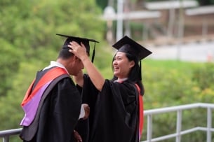 A student wearing a graduation robe and cap fixes a peer’s graduation cap