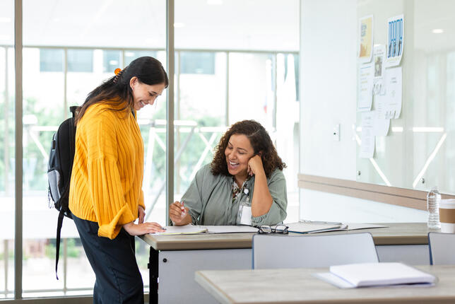Female student stands over a seated female professor who is helping her review materials on a desk in a brightly lit classroom.