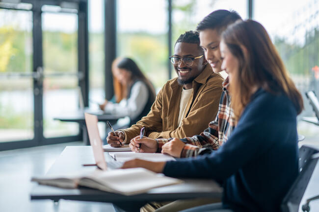 Three university students are seen sitting together in class as they work together on an assignment