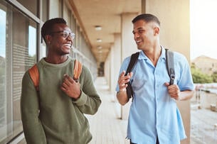 Two students wearing backpacks laugh while walking down a hallway at a college.