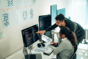 A man points to a computer screen as a woman examines charts.