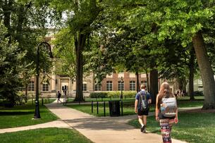 A photo of two college students wearing backpacks traversing a walking path on the Pennsylvania State University campus. The path is shaded with leafy trees and a large brick academic building can be seen in the background.