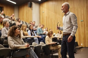 A senior male professor smiling at the front of a lecture hall full of engaged college students.