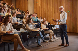 A male professor speaks at the front of a lecture hall filled with college students.