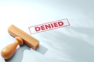 A wooden stamp lies atop a piece of paper on which it has seemingly just been used to stamp the word "DENIED" in red. 