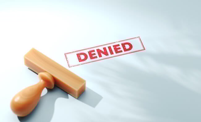 A wooden stamp lies atop a piece of paper on which it has seemingly just been used to stamp the word "DENIED" in red. 