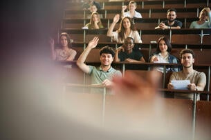 The outline of a professor can be seen facing a lecture hall with engaged college students, some with their hands raised.