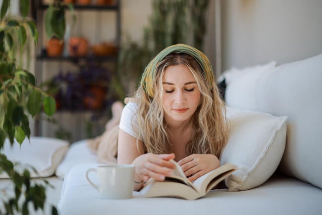 A photo of a young woman reading a book while lying face-down on a white couch in a room full of plants; a mug sits to the side of the open book in her hands.