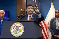 Illinois Governor JB Pritzker stands at a podium, pointing into the distance. He is a white man with dark hair, wearing a blue suit and red tie.
