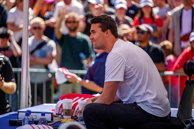 Charlie Kirk, in a white shirt, sits underneath a tent and behind a table with red and white hats and assorted coffee cups