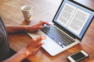 An author's hands rest on a laptop open to her book manuscript; a mug and cellphone sit off to the side. 
