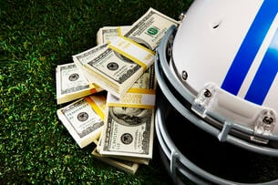 Stacks of $100 bills sit next to a football helmet atop a field.