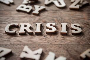 The word "crisis" is spelled out in wooden letters on top of a wooden table. Other random letters can be seen strewn about.