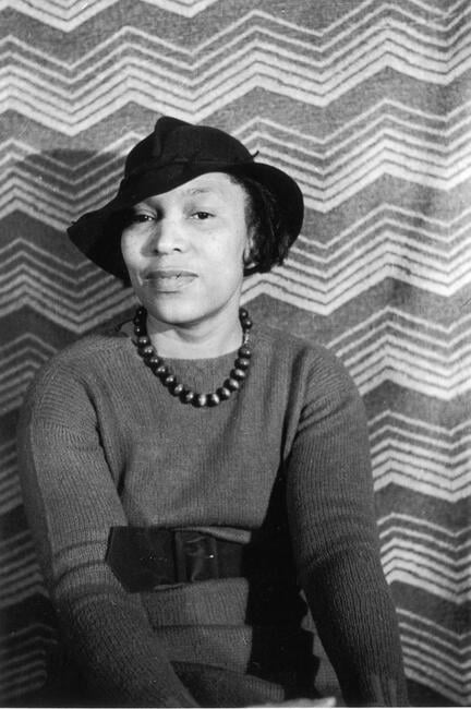 A portrait of author Zora Neale Hurston from around the 1940s. She wears a wide-brimmed hat and smiles while staring directly at the camera.