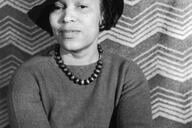 A portrait of author Zora Neale Hurston from around the 1940s. She wears a wide-brimmed hat and smiles while staring directly at the camera.