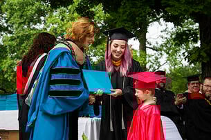 A mother and her young son, both dressed in graduation regalia, accept a diploma on stage from an administrator