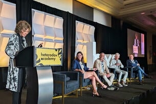 A photo of one person standing at a podium with the Heterodox Academy logo on it, to the left of four seated panelists.
