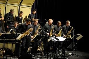 Five saxophone players stand to perform during a band concert. All students are clothed in black formal wear. 