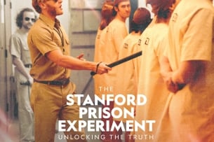 A poster for the 'Stanford Prison Experiment' limited series. A male student acting as a guard gestures with a baton toward a group of peers, dressed as prisoners, lined up and facing the other direction; text at the top states "Six days will last a lifetime." The poster also notes the series is streaming on Hulu and Disney+.