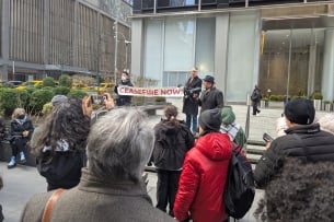 A photograph of a man speaking into a megaphone at a rally next to two people holding a banner reading "Ceasefire Now!"
