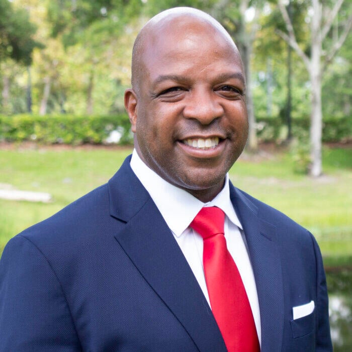 Sean Armstrong smiles for a headshot wearing a blue suit coat, red tie and white shirt. He is outside in a green area. 
