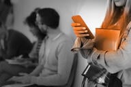 A student looks at a cell phone while standing in a hallway. The phone is highlighted in orange.
