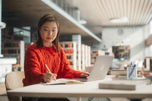 Student engaged in research with a laptop and notebook in a library. 