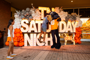 A student jumps to chestbump University of Tennessee at Knoxville mascot, Smokey, in front of a large balloon and light display that reads “TN Saturday Night”