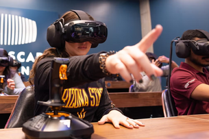 A classroom of students wearing black VR goggles, the student in the foreground has one hand outstretched in front of them